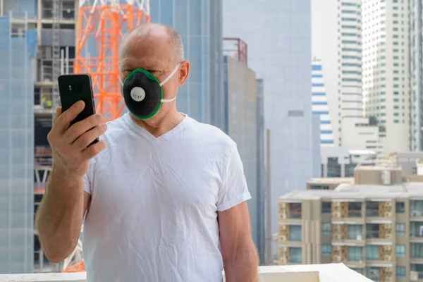 Старший чоловік використовує маску для обличчя, щоб захистити від забруднення смогу в місті, тримаючи мобільний телефон — стокове фото