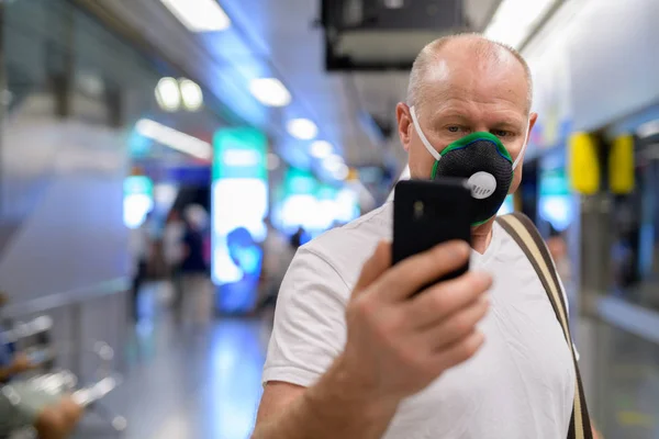 Старший мужчина использует маску для защиты от загрязнения смога в городе, держа мобильный телефон на вокзале — стоковое фото