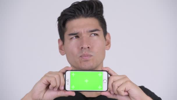 Gesicht eines glücklichen jungen gutaussehenden multiethnischen Mannes, der beim Vorzeigen seines Telefons denkt — Stockvideo
