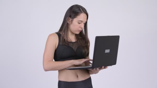 Joven feliz mujer multiétnica pensando mientras usa el ordenador portátil listo para el gimnasio — Vídeo de stock