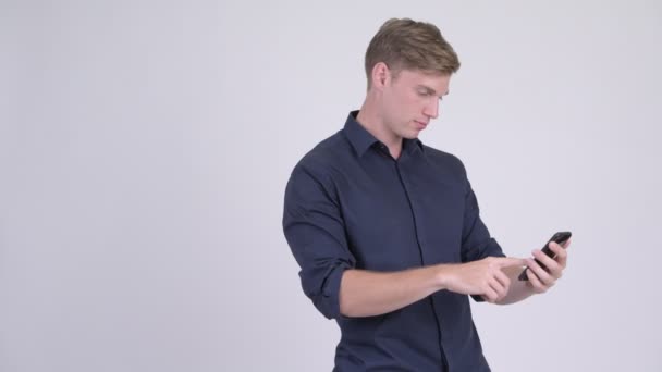 Молодой красивый блондин-бизнесмен пользуется телефоном и выглядит шокированным — стоковое видео