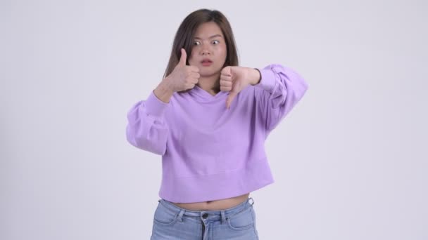 Joven mujer asiática confundida eligiendo entre los pulgares hacia arriba y hacia abajo — Vídeos de Stock
