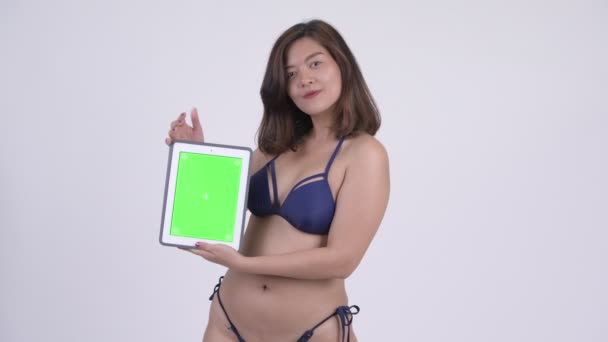 Счастливая молодая красивая азиатская туристка в бикини показывает цифровой планшет — стоковое видео