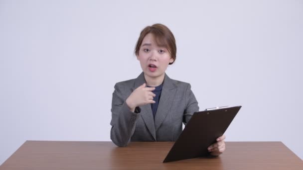 Young Asian businesswoman talking while holding clipboard at desk — Stock Video