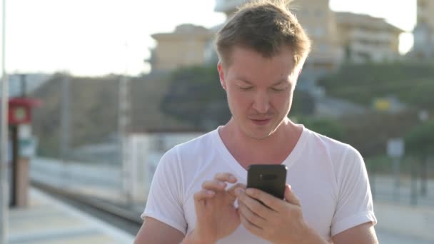 Joven turista feliz usando el teléfono mientras espera en la estación de tren — Vídeo de stock