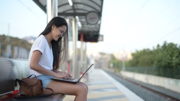 Joven feliz asiático turista mujer sentado y usando el ordenador portátil mientras espera el tren — Vídeos de Stock
