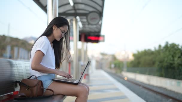 Νεαρό ζευγάρι πολυεθνικό χρησιμοποιώντας φορητό υπολογιστή μαζί, ενώ περιμένει στο σταθμό του τρένου — Αρχείο Βίντεο