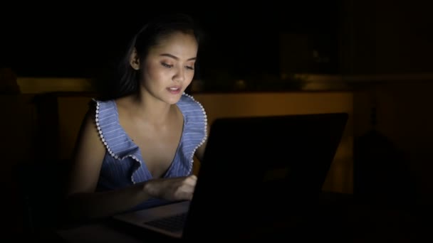 Молода красива азіатка посміхається під час використання ноутбука в темній кімнаті — стокове відео