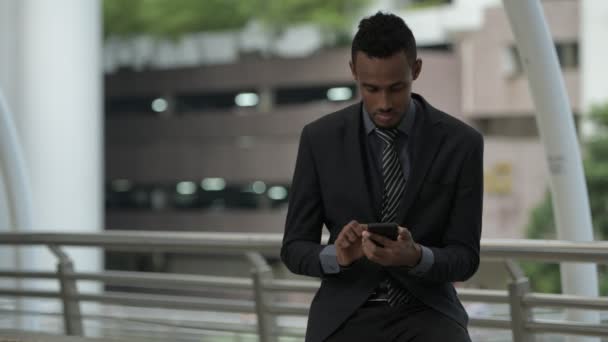 Jovem empresário africano pensando ao usar o telefone ao ar livre — Vídeo de Stock