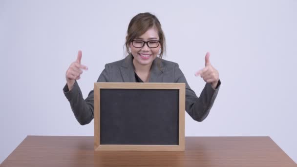 Unga glada asiatiska affärskvinna visar blackboard och ger tummen upp på skrivbord — Stockvideo