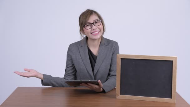 Unga glada asiatiska affärskvinna visar något med blackboard vid skrivbord — Stockvideo