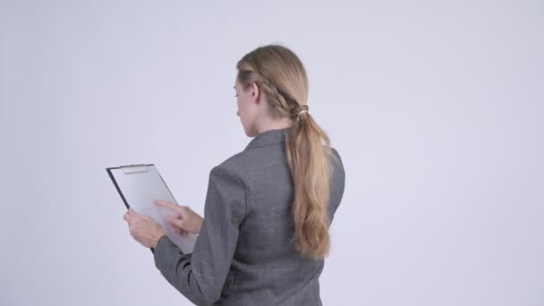 Vista trasera de la joven mujer de negocios rubia dirigiendo mientras lee en el portapapeles — Vídeos de Stock