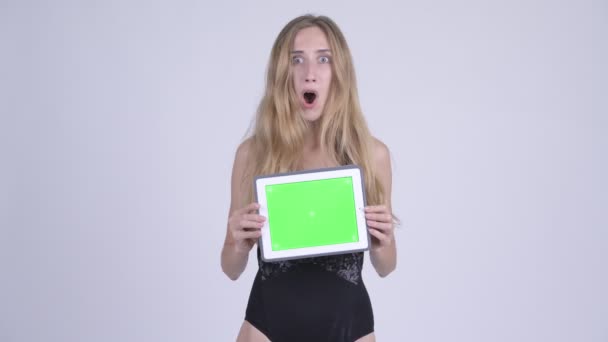 Mujer rubia joven feliz en traje de baño mostrando tableta digital y mirando sorprendido — Vídeos de Stock