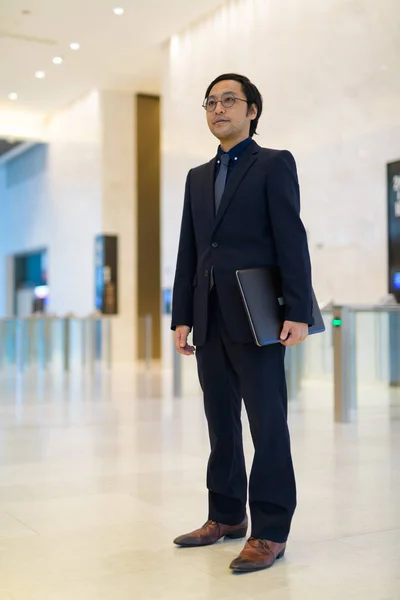 Cuerpo completo de hombre de negocios asiático sosteniendo portátil dentro del edificio de oficinas —  Fotos de Stock