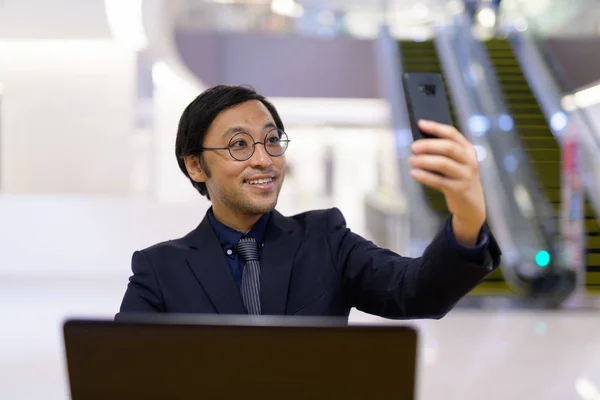 Happy Asian businessman using laptop and taking selfie inside the office building — Stock Photo, Image