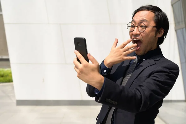 Empresário asiático feliz usando telefone e olhando surpreso ao ar livre — Fotografia de Stock