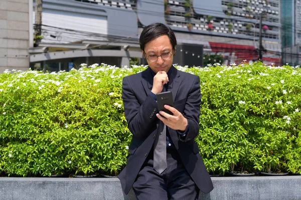 Hombre de negocios asiático pensando mientras usa el teléfono con la naturaleza en la ciudad —  Fotos de Stock