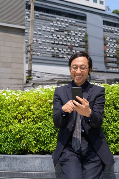 Feliz hombre de negocios asiático riendo mientras usa el teléfono con la naturaleza en la ciudad —  Fotos de Stock