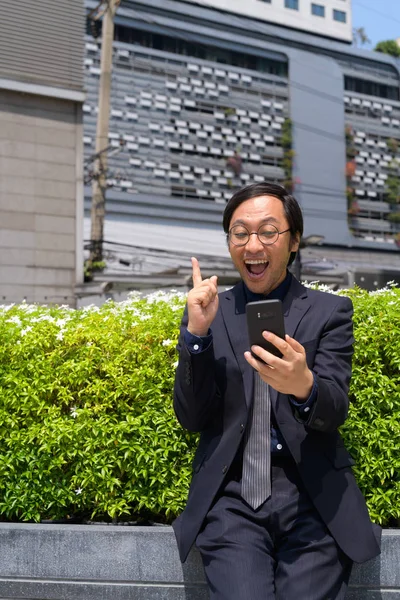 Feliz asiático hombre de negocios apuntando hacia arriba mientras se utiliza el teléfono en la ciudad al aire libre —  Fotos de Stock