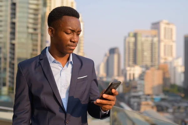 Joven hombre de negocios africano guapo usando el teléfono contra la vista de la ciudad — Foto de Stock