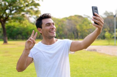 Mutlu genç yakışıklı İspanyol adam selfie barış işareti ile parkta alarak