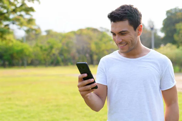 Heureux jeune homme hispanique beau utilisant le téléphone dans le parc à l'extérieur — Photo