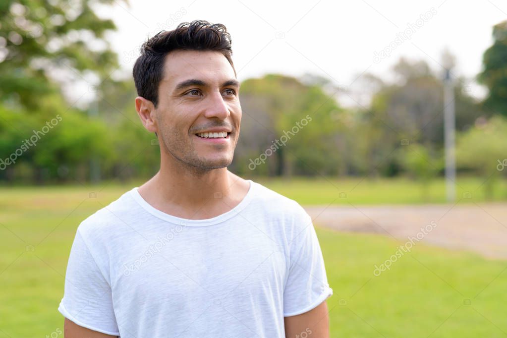 Face of happy young handsome Hispanic man thinking at the park