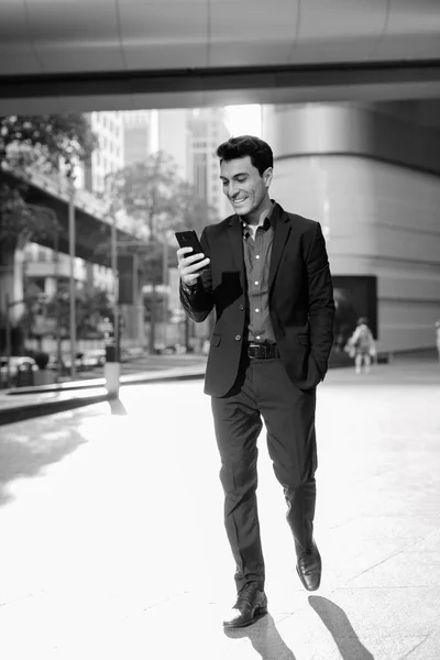 Jeune homme d'affaires hispanique heureux utilisant le téléphone à l'extérieur de l'immeuble de bureaux en noir et blanc — Photo