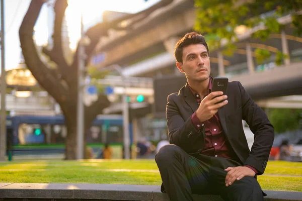 Young thoughtful Hispanic businessman holding phone and sitting with nature in city — Stock Photo, Image