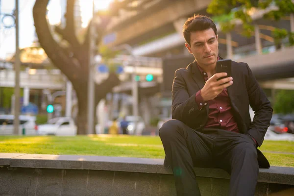 Jeune homme d'affaires hispanique beau utilisant le téléphone et assis avec la nature en ville — Photo