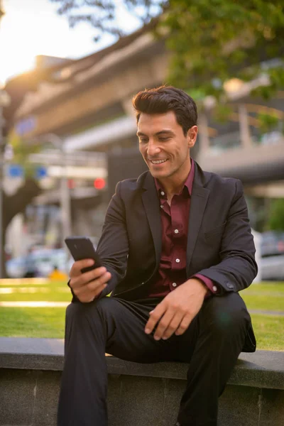 Heureux bel homme d'affaires hispanique utilisant le téléphone et assis avec la nature en ville — Photo