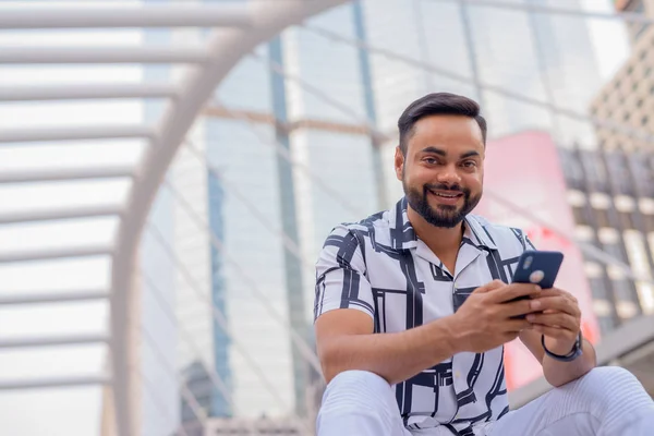 Heureux jeune homme indien barbu avec téléphone assis dans la ville en plein air — Photo