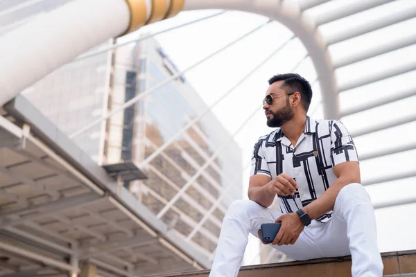 Young bearded Indian man with sunglasses sitting in the city outdoors