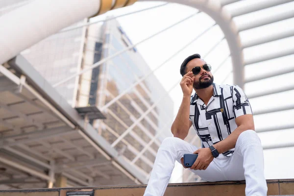 Jeune homme indien barbu avec des lunettes de soleil assis en ville à l'extérieur — Photo