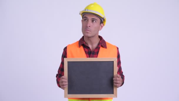 Joven feliz trabajador de la construcción hispano pensando mientras sostiene pizarra — Vídeos de Stock