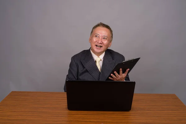 Mogen asiatisk affärsman sitter med träbord mot grått — Stockfoto
