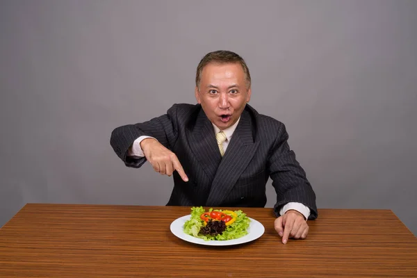 Volwassen Aziatische zakenman zitten met houten tafel tegen grijs — Stockfoto