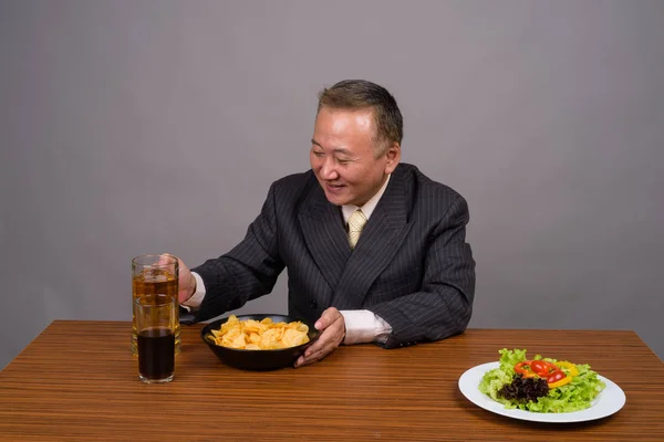 Volwassen Aziatische zakenman zitten met houten tafel tegen grijs — Stockfoto