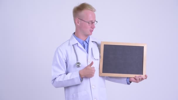 Happy young blonde man doctor holding blackboard and giving thumbs up — Stock Video