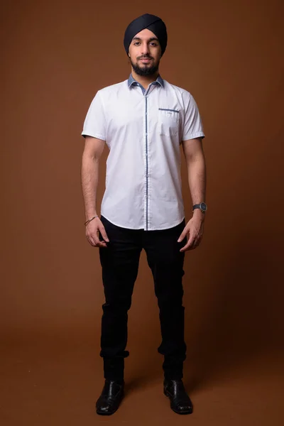 Young bearded Indian Sikh man wearing turban against brown backg