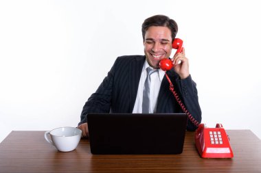 Studio shot of young happy Persian businessman smiling and talking on old telephone while using laptop with coffee cup on wooden table against white background