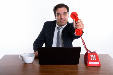 Studio shot of young Persian businessman giving old telephone with laptop and coffee cup on wooden table against white background