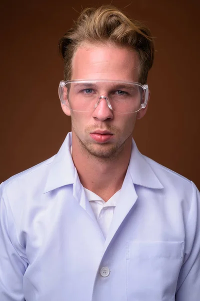 工作室拍摄的年轻英俊男子医生戴防护眼镜对棕色背景 — 图库照片