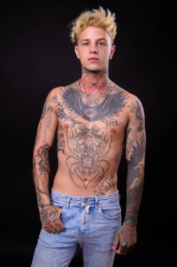 Studio shot of young handsome rebellious man with blond hair and body tattoos shirtless against black background