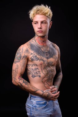 Studio shot of young handsome rebellious man with blond hair and body tattoos shirtless against black background
