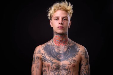 Studio shot of young handsome rebellious man with blond hair and body tattoos shirtless against black background