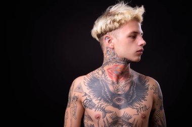 Studio shot of young handsome rebellious man with blond hair and body tattoos shirtless against black background