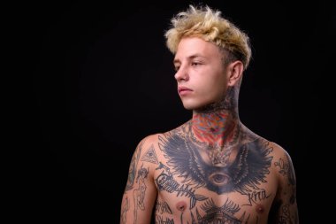 Studio shot of young handsome rebellious man with blond hair and body tattoos shirtless against black background