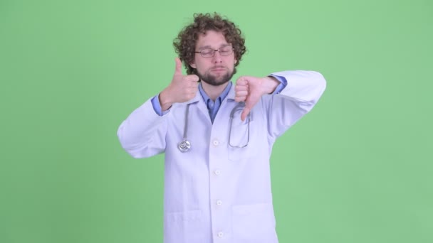 Запутавшийся молодой бородатый врач выбирает между большим и большим пальцами вниз — стоковое видео