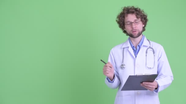 Happy young bearded man doctor presenting something while writing on clipboard — Stock Video